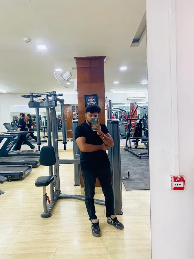 A gym floor reflected in a wall mirror: a row of treadmills, a cable machine, and a person in gym clothes standing among the equipment.